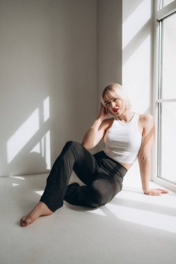 Beyaz bluzlu ve siyah pantolonlu sarışın bir kadın pencerenin kenarında oturur ve doğal ışıkla yıkanır. Kendine güveni ve tarzı var..