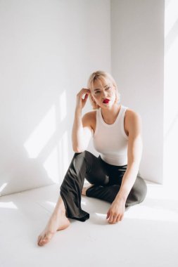 Kırmızı rujlu ve büyük halka küpeli sarışın bir kadın, parlak, minimalist bir odada, beyaz ve siyah bir pantolonla yerde oturuyor..