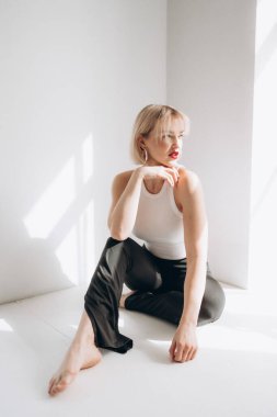 Kırmızı rujlu ve büyük halka küpeli sarışın bir kadın, parlak, minimalist bir odada, beyaz ve siyah bir pantolonla yerde oturuyor..