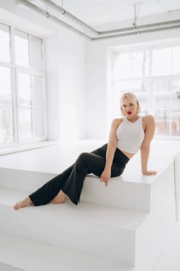 Beyaz bluzlu, kırmızı rujlu ve siyah pantolonlu sarışın bir kadın parlak, minimalist bir odada beyaz basamaklarda çıplak ayakla poz veriyor. Boşluğu kopyala.