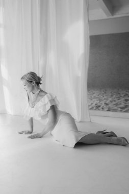 Sarışın, kısa saçlı, kırmızı rujlu beyaz bir elbise ve inci kolyeyle yere poz veren zarif bir kadının siyah beyaz fotoğrafı. Boşluğu kopyala.