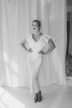 Sarışın, kısa saçlı, kırmızı rujlu beyaz elbiseli ve inci kolyeli zarif bir kadının beyaz arka planda durduğu siyah beyaz bir fotoğraf..