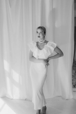 Sarışın, kısa saçlı, kırmızı rujlu beyaz elbiseli ve inci kolyeli zarif bir kadının beyaz arka planda durduğu siyah beyaz bir fotoğraf..