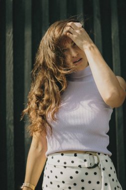 Uzun saçlı, lavanta rengi bluzlu ve benekli etekli bir kadın, kafasına dokunuyor, doğal ışıkta koyu bir arka planda duruyor..