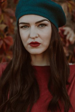Arka planda sonbahar yeşilliği olan, deniz mavisi bereli ve kestane rengi bluzlu bir kadının yakın plan portresi..