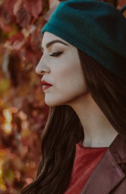 Deniz mavisi bereli ve kestane rengi deri ceketli bir kadının yan profili sonbahar yeşilliklerinin arka planına karşı..