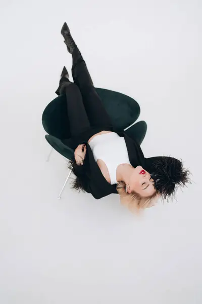 Stock image Woman in stylish black blazer and high heels lying upside down on a chair, showcasing a modern and chic fashion pose.