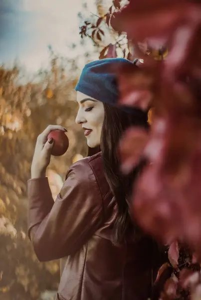 stock image Woman in teal beret and maroon leather jacket holding an apple, surrounded by autumn foliage.