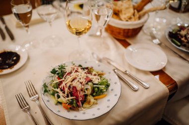 Riga, Letonya 20 Mart 2024 - Taze salata, şarap bardağı ve ekmek sepetli bir yemek masası. Zarif çatal bıçak ve cam takımlar düzgün bir şekilde dizilmiş. Boşluğu kopyala.