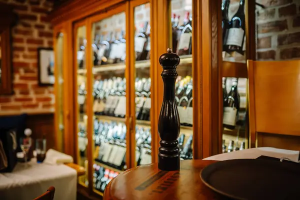 stock image Riga, Latvia March 20, 2024 - Close-up of a black pepper mill on a wooden table in a cozy restaurant, with a background of wine bottles in illuminated wooden cabinets.