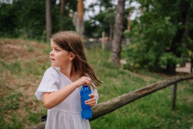 Riga, Letonya - 19 Temmuz 2024 - Elinde mavi bir şişe tutan beyaz elbiseli genç bir kız tahta bir çitin yanında duruyor, arka planda ağaçlarla uzaklığa bakıyor.