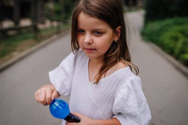 Riga, Letonya - 19 Temmuz 2024 - Beyaz elbiseli genç kız park yolunda mavi bir oyuncak tutuyor, arkasında ağaçlar ve yeşillik var.