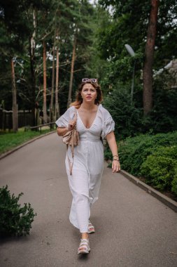 Riga, Letonya - 19 Temmuz 2024 - Beyaz elbiseli bir kadın park yolunda yürüyor, arkasında ağaçlar ve yeşillik olan bej bir çanta taşıyor.