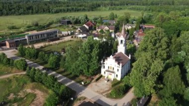 Lielvarde, Letonya - 20 Haziran 2024 - Yeşil tarlalarla çevrili güneşli bir günde kilise, evler, ağaçlar ve insanların bulunduğu kırsal bir köyün havadan görünüşü.