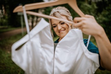 Cesis, Letonya - 12 Temmuz 2024 - Tacını takmış gülümseyen bir kadın, açık havada asılı beyaz bir elbiseyi yakından inceliyor..