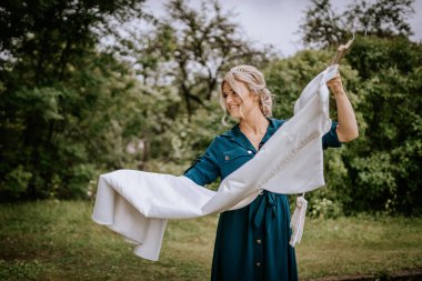 Cesis, Letonya - 12 Temmuz 2024 - Yeşil elbiseli gülümseyen bir kadın bulutlu bir günde yemyeşil bir hava ile çevrili bir askıda beyaz bir elbise tutuyor..