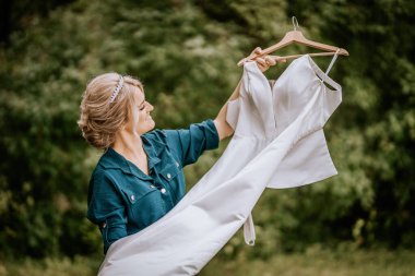 Cesis, Letonya - 12 Temmuz 2024 - Tarzlı saçlı gülümseyen bir kadın, açık havada yeşil yapraklı arka planda beyaz bir elbise tutuyor..