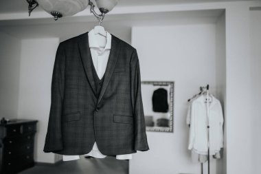Cesis, Letonya - 12 Temmuz 2024 - Askıda koyu renk bir takım elbise ve beyaz gömlek, ayna ve giysi askısı da dahil olmak üzere bulanık arka planı olan bir odada sergileniyor. Görüntü siyah beyaz..