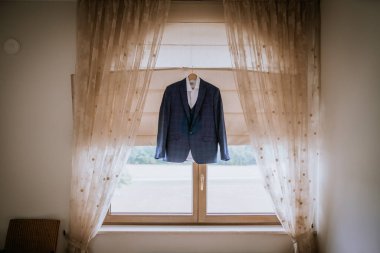 Cesis, Letonya - 12 Temmuz 2024 - Bir askı üzerinde koyu renk bir ceket ve beyaz gömlek bir pencerenin önünde sergileniyor..