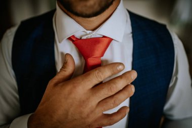 Cesis, Letonya - 12 Temmuz 2024 - Kırmızı kravatını düzelten, beyaz gömlek ve mavi yelek giyen, eline odaklanan ve kravat düğümlü bir adamın yakın çekimi.