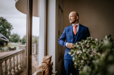 Cesis, Letonya - 12 Temmuz 2024 - Mavi takım elbiseli ve kırmızı kravatlı bir adam büyük bir pencerenin yanında duruyor, kendine güvenen bir ifadeyle dışarı bakıyor, yeşil ışıklı bir ev ortamında.