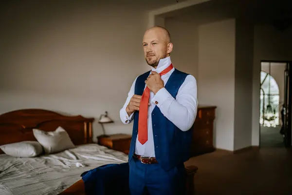 stock image Cesis, Latvia - July 12, 2024 - A man in a blue vest and white shirt adjusts his red tie in a bedroom, preparing for a formal event. A bed and dresser are visible in the background.