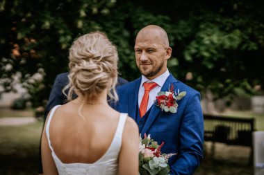 Cesis, Letonya - 12 Temmuz 2024 - Kırmızı kravatlı ve yaka çiçekli bir damat açık hava düğün törenlerinde geline sevgiyle bakıyor.