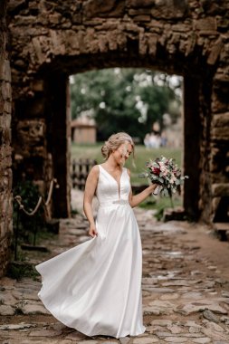 Cesis, Letonya - 12 Temmuz 2024 - Dalgalı beyaz elbiseli neşeli bir gelin, çiçeğiyle taş kemer yolunda poz verir, düğün gününde mutluluk saçar.