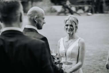 Cesis, Letonya - 12 Temmuz 2024 - Gelin ve damat açık havada yapılan bir düğün sırasında el ele tutuşuyorlar, gelin nazikçe gülümsüyor. Siyah ve beyaz resim.