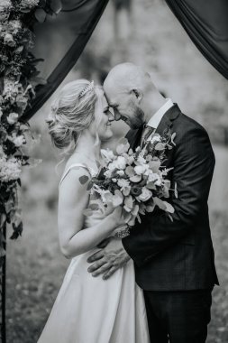 Cesis, Latvia - July 12, 2024 - A bride and groom share an intimate moment, forehead to forehead, while holding a bouquet. The image is captured in black and white. clipart