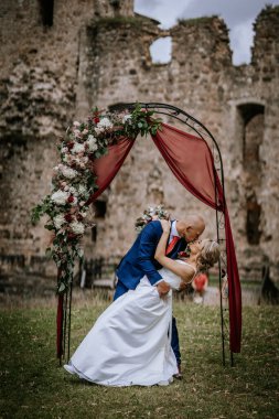 Cesis, Latvia - July 12, 2024 - A groom romantically dips and kisses his bride under a decorated floral arch with an ancient stone building in the background during their wedding. clipart