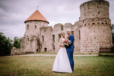 Cesis, Letonya - 12 Temmuz 2024 - Bir gelin ve damat, gelin bir buket tutarken ve damat onu kucaklarken antik taş bir kalenin önünde poz veriyorlar. Gökyüzü kapalı..