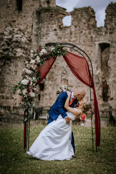 stock image Cesis, Latvia - July 12, 2024 - A groom romantically dips and kisses his bride under a decorated floral arch with an ancient stone building in the background during their wedding.