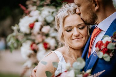 Cesis, Letonya - 12 Temmuz 2024 - Bir gelin damadıyla hassas bir anın tadını çıkarır, o da alnından öper. Etrafları çiçekler ve yeşilliklerle çevrili. Romantik bir ortam yaratıyorlar..