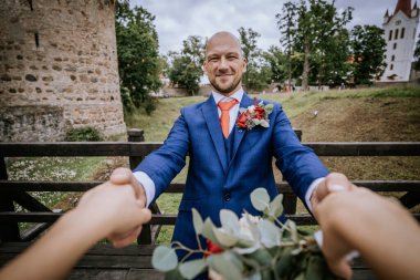 Cesis, Letonya - 12 Temmuz 2024 - Kırmızı yaka çiçekli mavi takım elbiseli bir damat, arka planda tarihi harabeler ve yeşilliklerle gelinin elini tutarken gülümsüyor.