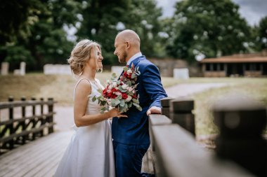 Cesis, Letonya - 12 Temmuz 2024 - Gelin ve damat ahşap bir köprüde, gelin bir buket tutuyor ve birbirlerine sevgiyle gülümsüyorlar..