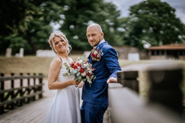 Cesis, Letonya - 12 Temmuz 2024 - Bir gelin ve damat ahşap bir köprüde poz veriyor, ikisi de sıcak bir şekilde gülümsüyor. Damat korkuluklara yaslanırken gelin bir buket tutuyor..