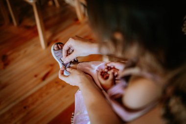 Valmiera, Letonya - 15 Temmuz 2024 - Çocuk pembe bir elbisenin içinde, arka planda ahşap döşeme ile otururken genç bir çocuğun sandalet bağını bağlayan bir yetişkinin yakın çekimi.