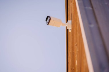 Ahşap bir duvara monte edilmiş bir güvenlik kamerası, çevreyi izlemek için açılı, arka plan olarak açık mavi gökyüzü ve geniş kopya alanı ile.