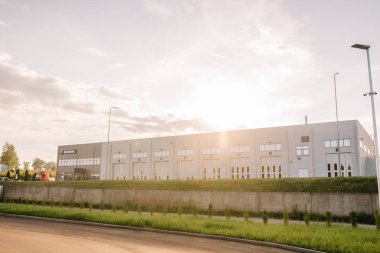 Valmiera, Letonya - 8 Ağustos 2024 - Günbatımında birçok büyük kapısı ve penceresi olan modern endüstriyel bir bina, yumuşak güneş ışığıyla manzarayı aydınlatıyor. 
