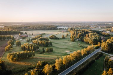 Valmiera, Letonya - 15 Ağustos 2024 - Güneş doğarken uzak bir kasaba olan ağaçlar ve tarlalarla çevrili bir golf sahasının havadan görünüşü. Boşluğu kopyala.