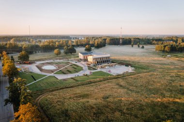 Valmiera, Letonya - 15 Ağustos 2024 - Güneşin doğuşuyla birlikte kırsal bir bölgede manzaralı bir bina ve ağaçlarla çevrili modern bir bina. Boşluğu kopyala.