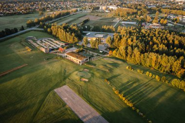 Valmiera, Letonya - 15 Ağustos 2024 - Golf sahasının havadan görünüşü, golf sahaları, bir kulüp evi, çevre ağaçlar ve gün doğumunda yakınlardaki kasaba binaları. Boşluğu kopyala.