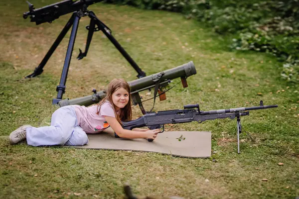 stock image A young girl lies on the grass, holding a large machine gun with a smile, near military equipment during an outdoor event.