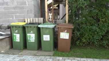 Valmiera, Letonya - 28 Ağustos 2024 - Bir binanın ve yeşilliklerin yanına yerleştirilmiş yeşil ve kahverengi kapaklı bir sıra geri dönüşüm ve atık kutuları.