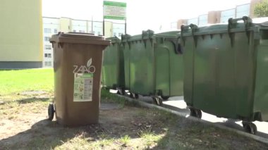 Valmiera, Letonya - 28 Ağustos 2024 - Güneşli bir günde apartman binalarının yakınındaki bir açık alanda yeşil atık kutularının önünde kahverengi bir geri dönüşüm kutusu duruyor.