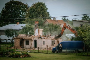 Valmiera, Letonya - 16 Ağustos 2024 - Ağaçlar ve diğer evlerle çevrili bir yerleşim bölgesinde mavi kamyonetli bir kazıcı tarafından bir ev yıkılıyor.