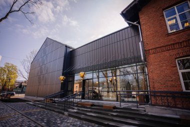 Valmiera, Letonya - 26 Nisan 2024 - Camdan kapıları ve basamakları olan modern bir bina, cephede altın ödüllü süslemeler, kentsel bir ortamda tuğla bir yapının yanında.
