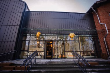 Valmiera, Letonya - 26 Nisan 2024 - Camdan kapıları ve basamakları olan, altın kupa dekorasyonlarıyla süslenmiş, içeriden yansıyan ağaçlar ve ışıkları olan modern bir bina girişi.