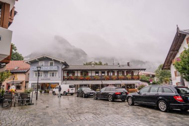 Oberammergau, Germany - September 14, 2024 - A rainy European street with parked cars, shops, and restaurants, framed by fog-covered mountains in the background, creating a cozy village atmosphere. clipart